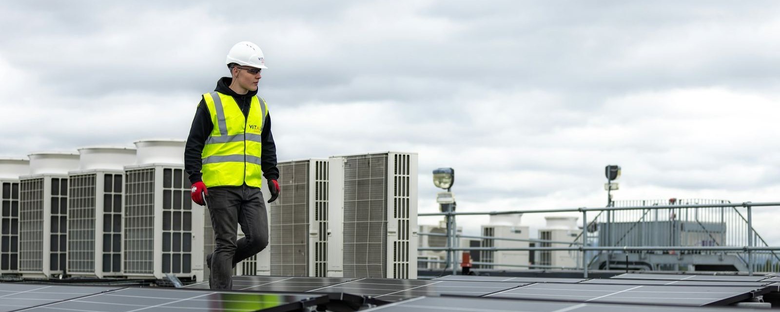 Technician in a hard hat and hi-vis vest walking across a roof past the outdoor units