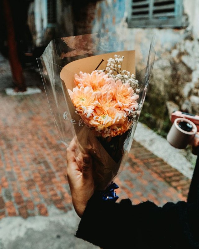 Ramo de flores anaranjadas en papel kraft y celofán, en la mano, por una calle de adoquines