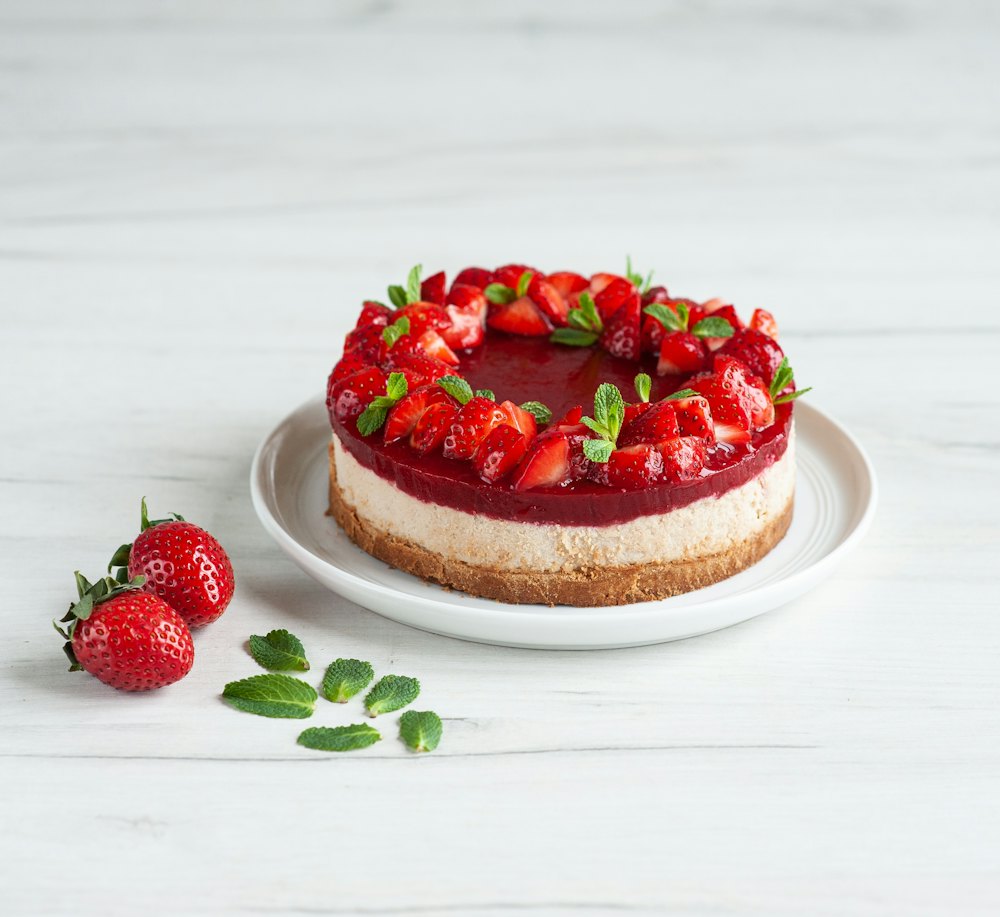 A whole strawberry cheesecake on a white plate, with strawberries and mint leaves beside it