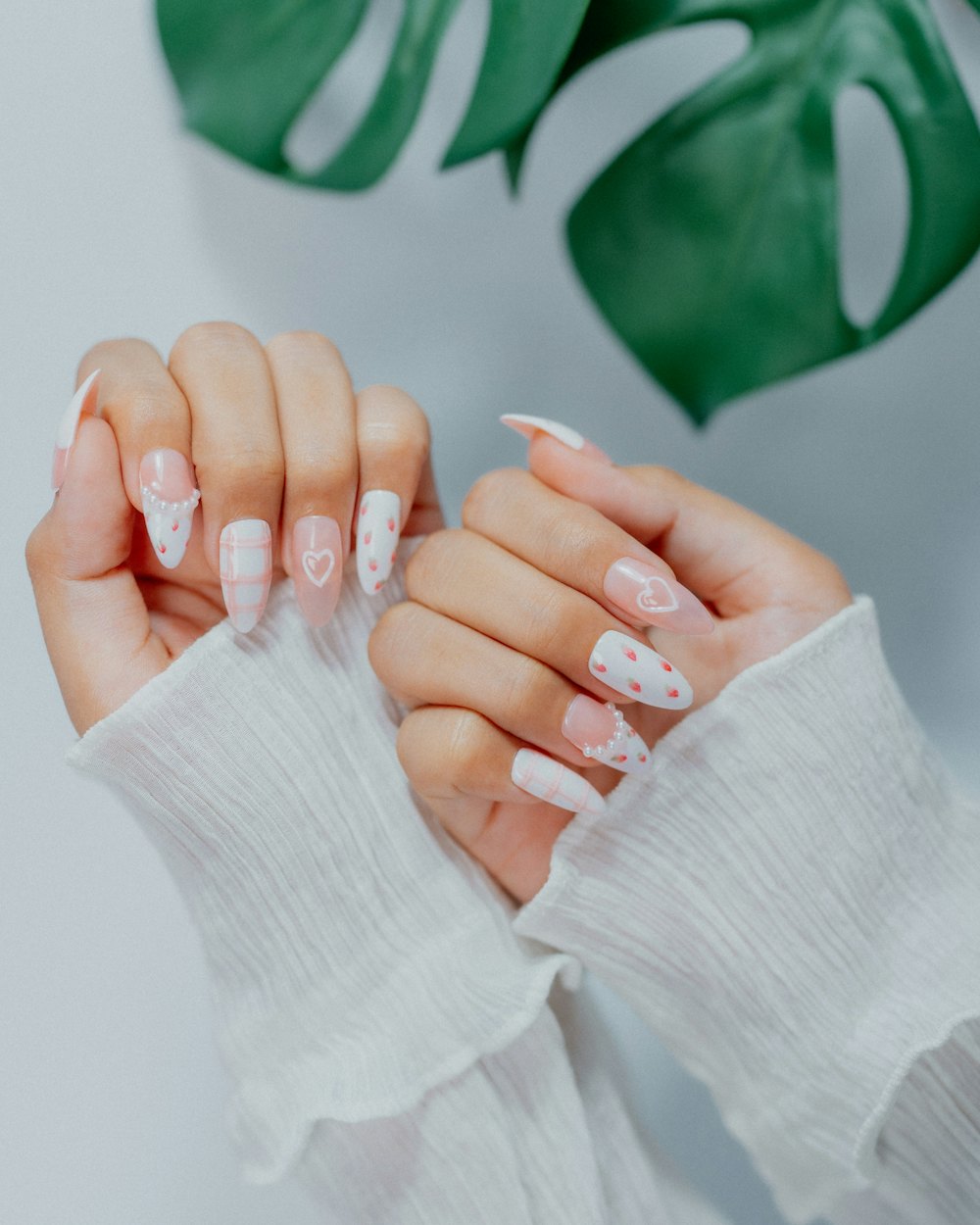 Manos con uñas largas en blanco y pequeños dibujos, junto a una hoja de monstera